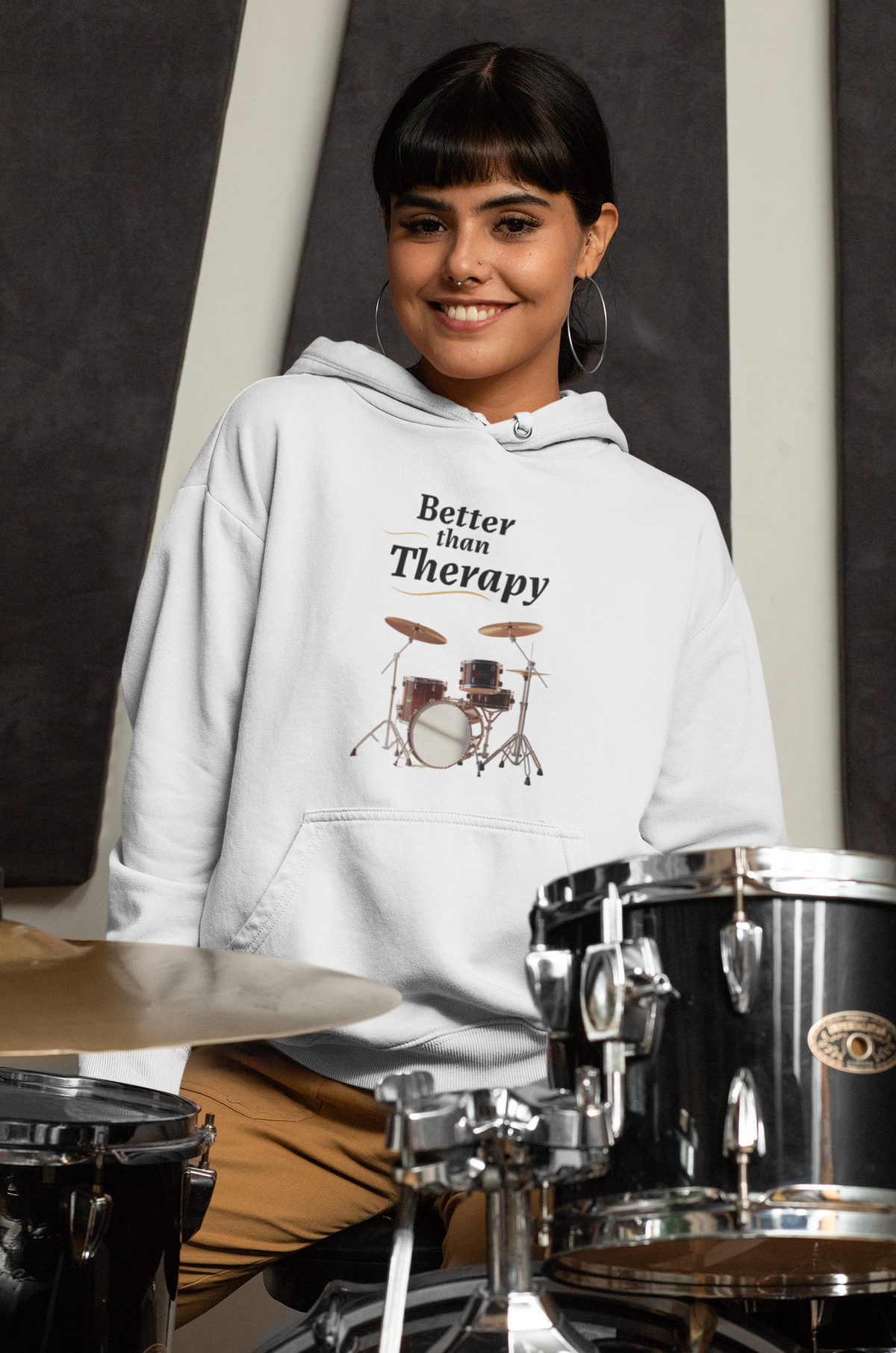 Person wearing a hoodie with 'Better than Therapy' text and drum design, standing in front of drums.